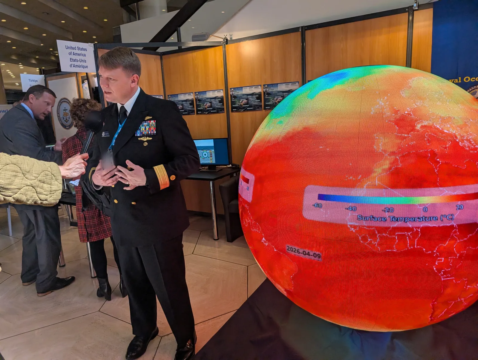 A naval officer being interviewed beside the Ayni sphere, which is showing global surface temperature with its colour legend and date visible on the display