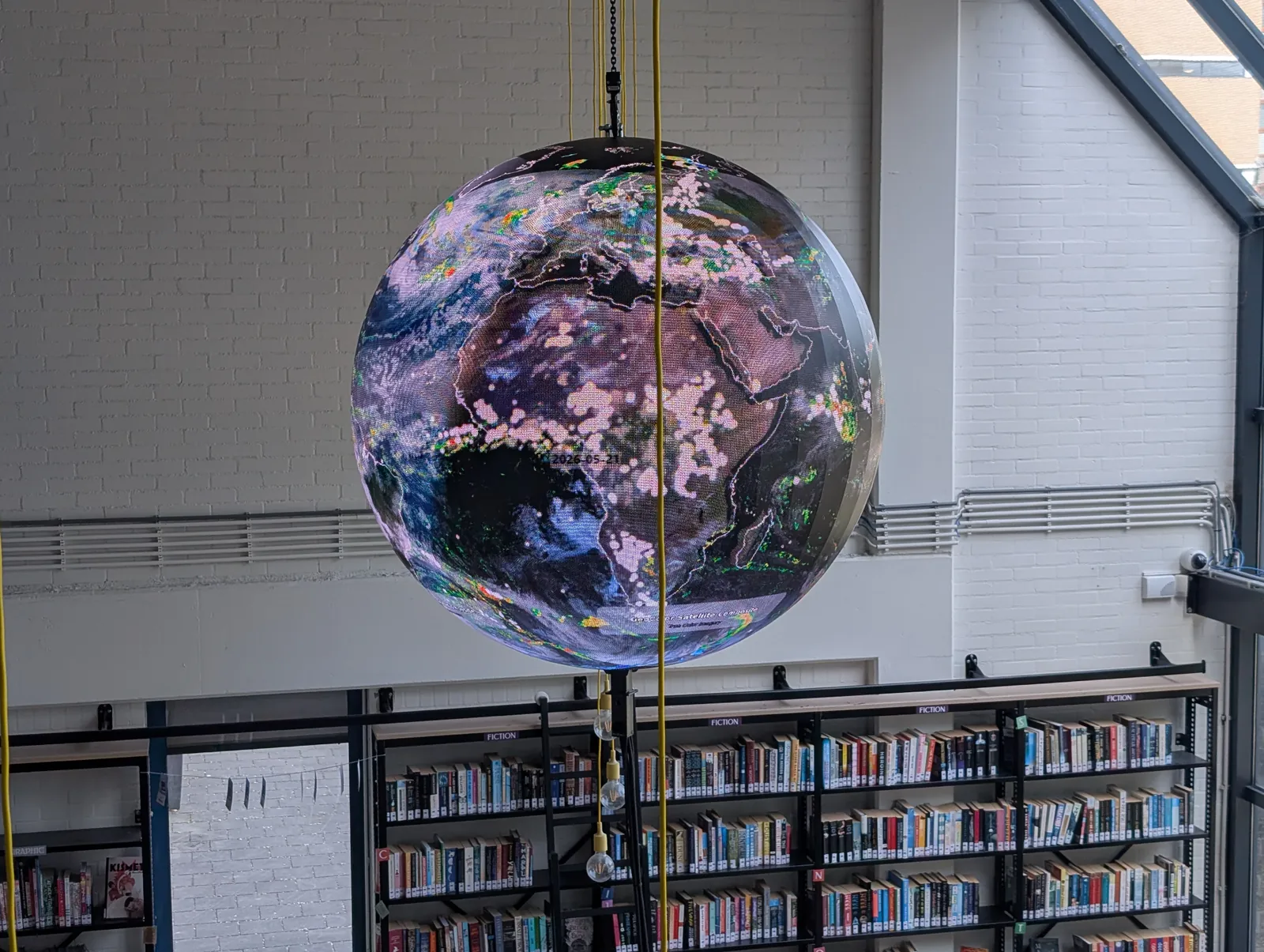 An Ayni sphere suspended above the shelves of a school library, showing a dated global satellite composite